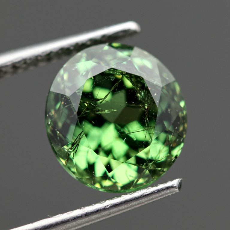 Primer plano de un granate Demantoide verde redondo facetado, con inclusiones fibrosas internas visibles, sostenido por pinzas.